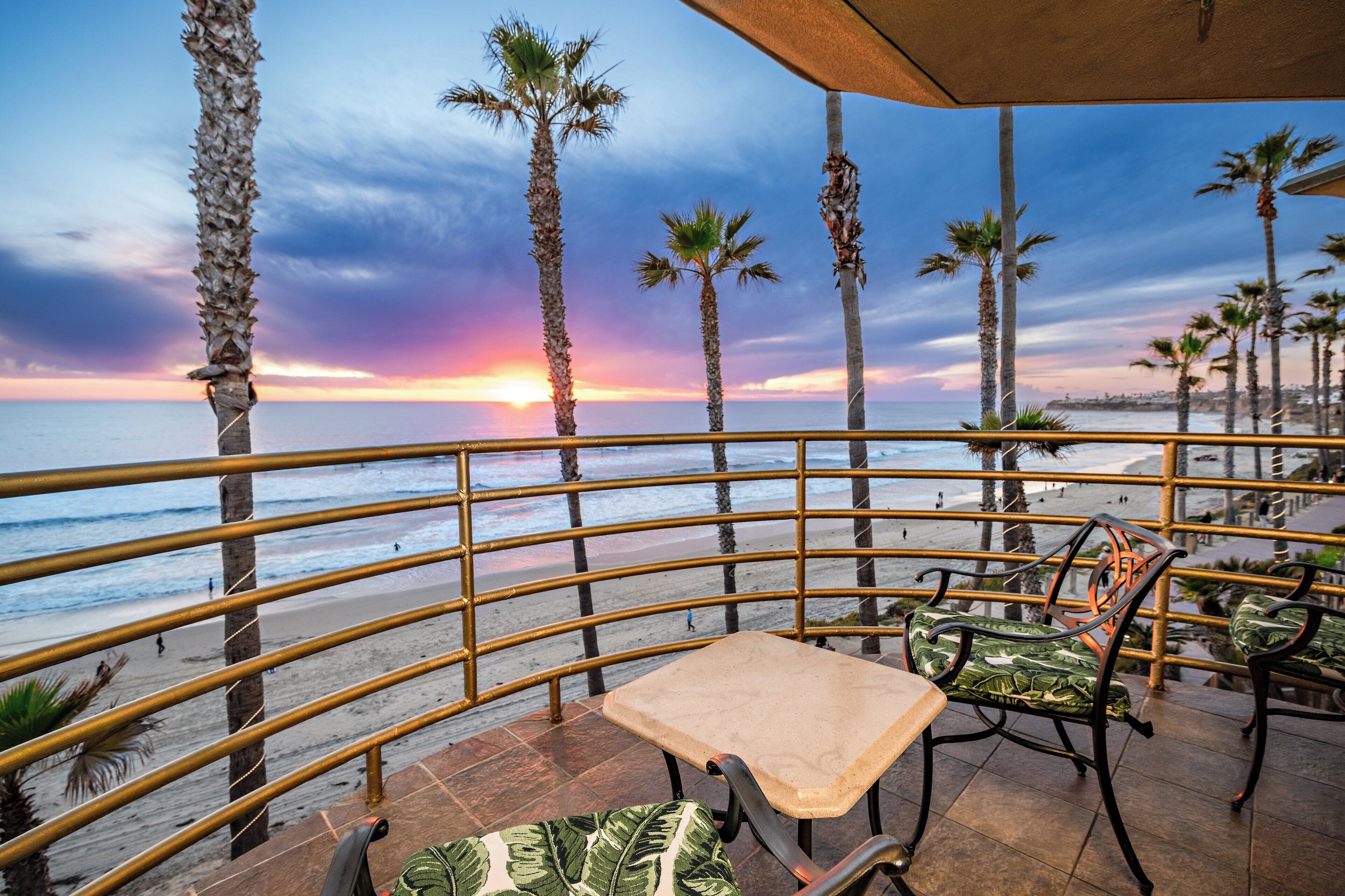 A terrace with a view of the beach and sunset, featuring palm trees, a railing, and outdoor seating.