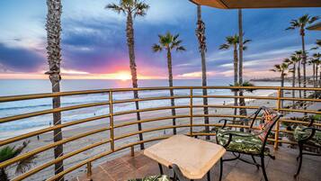 A terrace with a view of the beach and sunset, featuring palm trees, a railing, and outdoor seating.