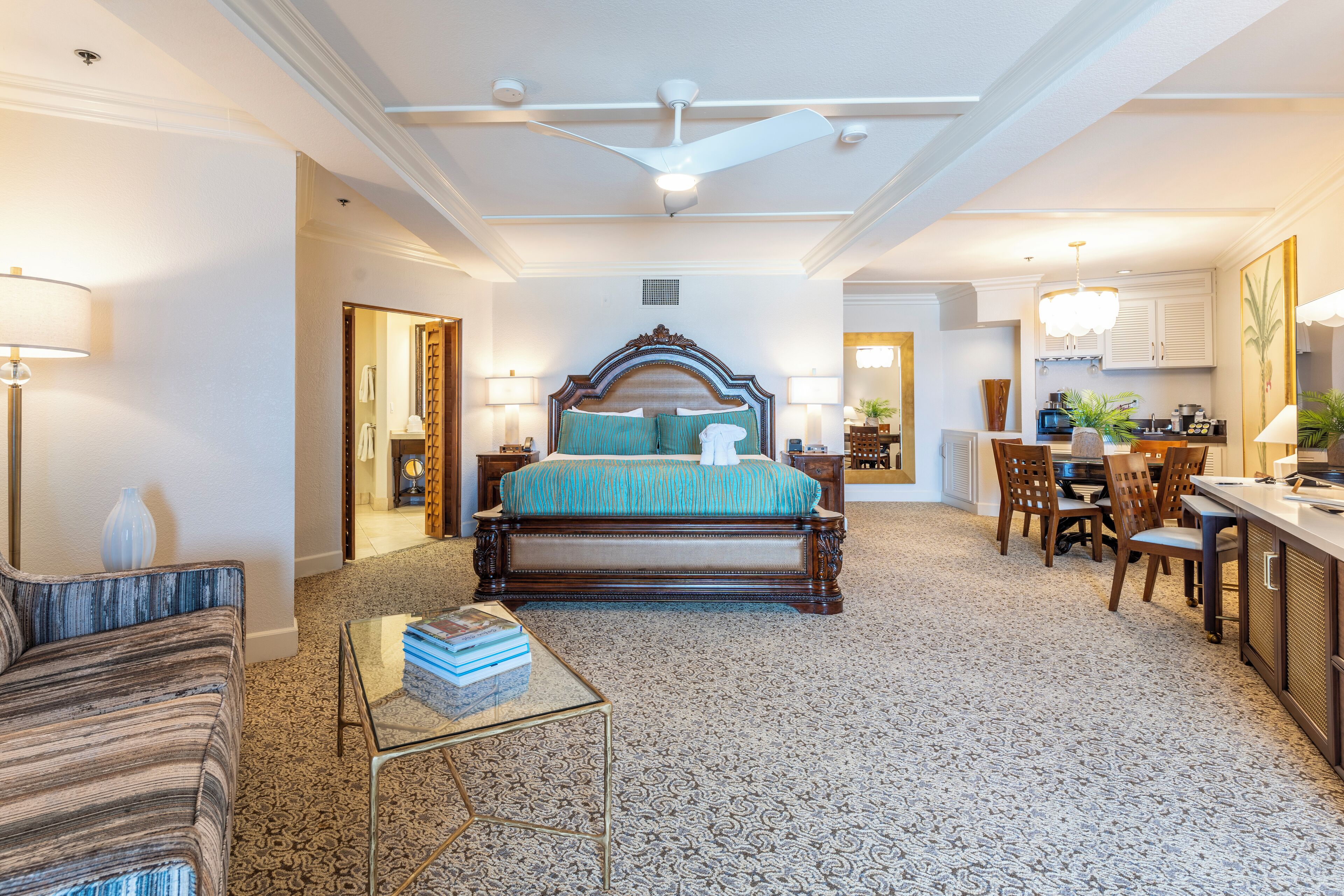 A spacious hotel room with a large bed, a sitting area with a sofa and a small table, a kitchenette, and a bathroom visible through an open door.