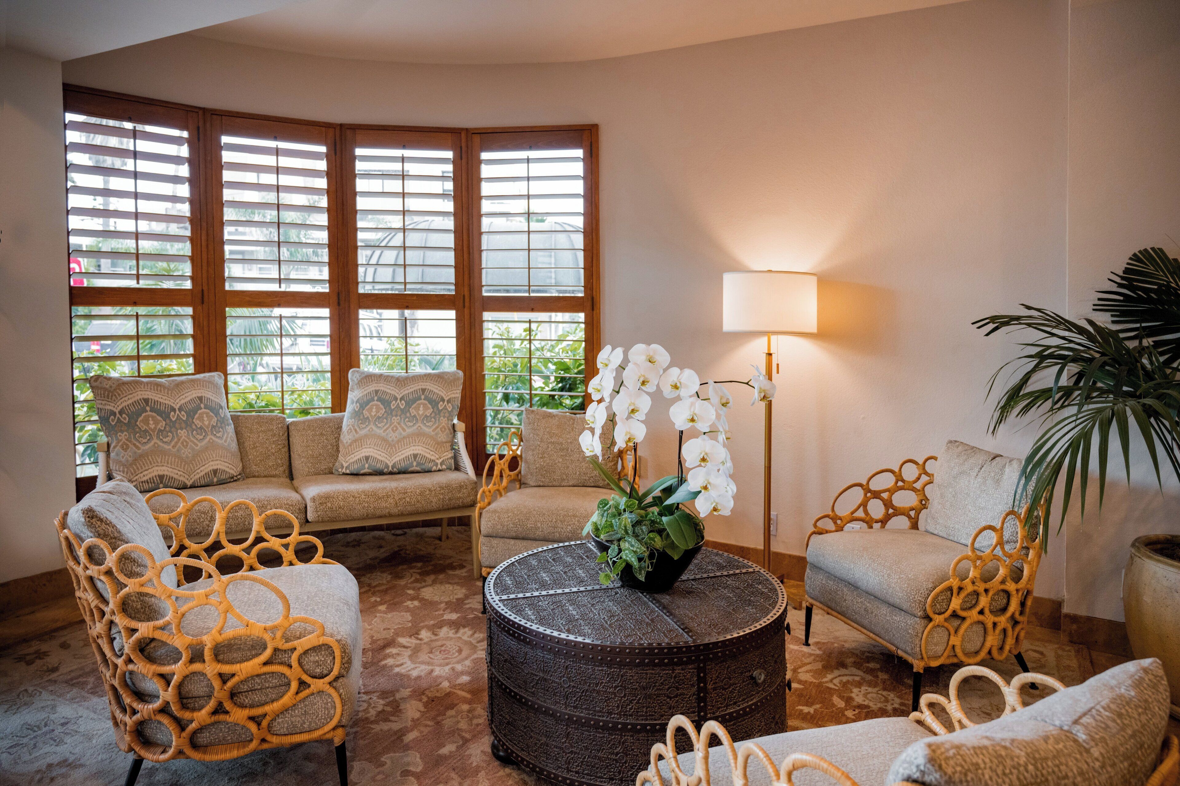 Un salon chaleureux avec un canapé, deux fauteuils, un pouf rond et une lampe sur pied. De grandes fenêtres équipées de stores laissent entrer la lumière naturelle et illuminent l’espace.
