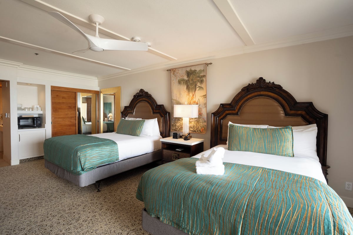 A hotel room with two beds, a ceiling fan, a painting, and a microwave.