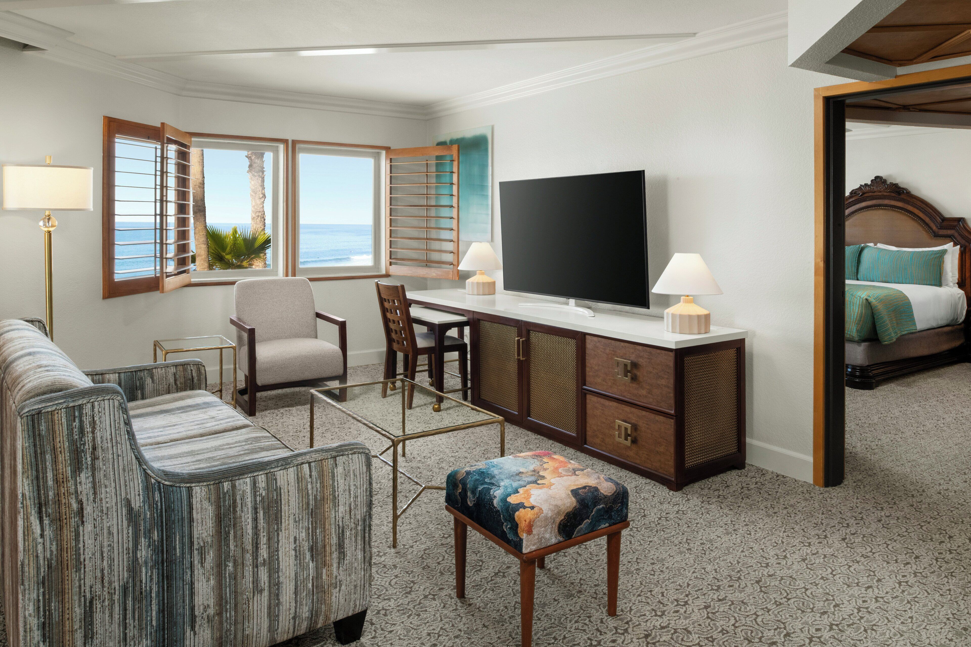 A hotel room with a large flat-screen TV, a desk with a chair, a sofa, a small ottoman, and a bed with a wooden headboard.