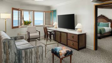 A hotel room with a large flat-screen TV, a desk with a chair, a sofa, a small ottoman, and a bed with a wooden headboard.