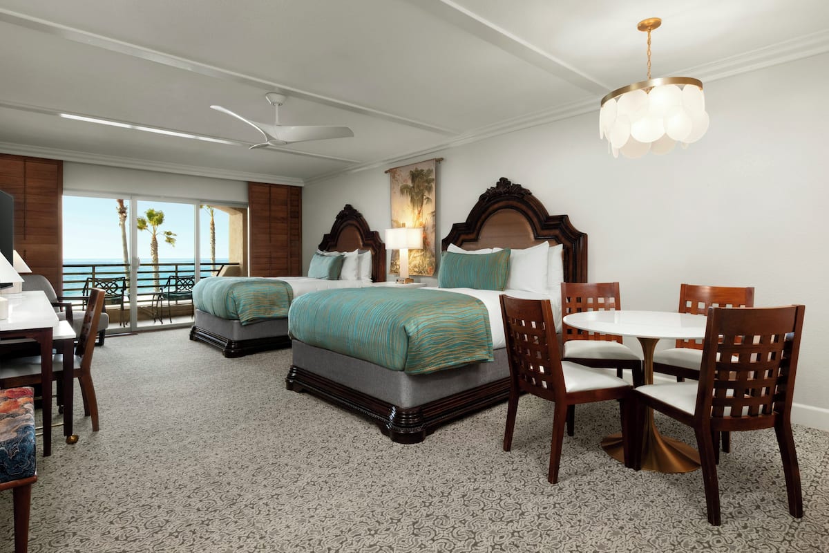 A hotel room with two beds, a dining table, chairs, a ceiling fan, and a view of the ocean.