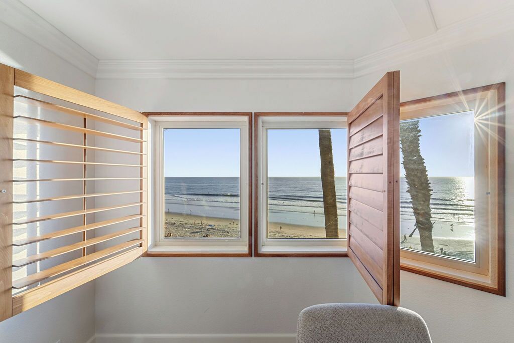 A room with a large bay window overlooking a beach, a wooden slatted window blind, and a gray chair.