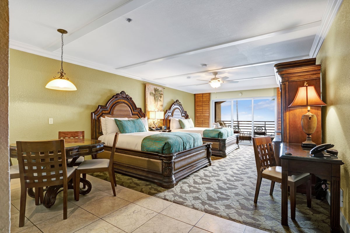A hotel room with a bed, desk, chairs, and a balcony with a view.
