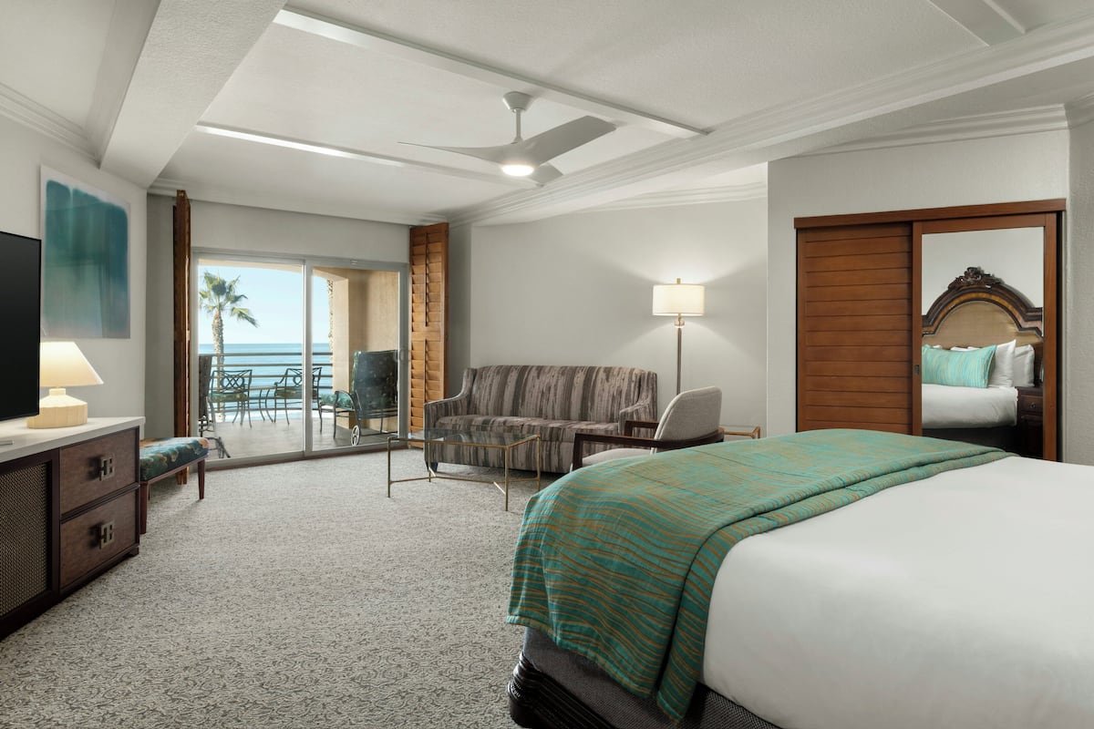 A hotel room with a bed, a desk with a lamp, a chair, a sofa, a TV, and a view of the ocean.