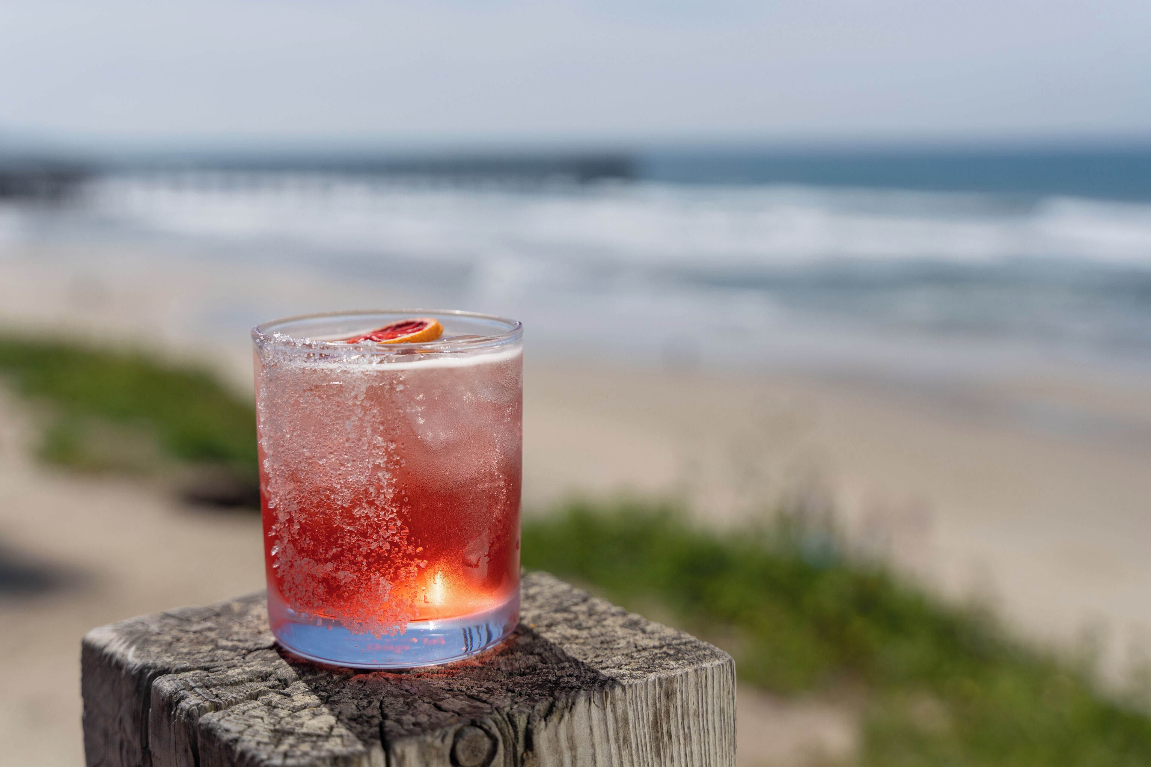 A glass of iced beverage with a slice of fruit on a wooden post by the beach.