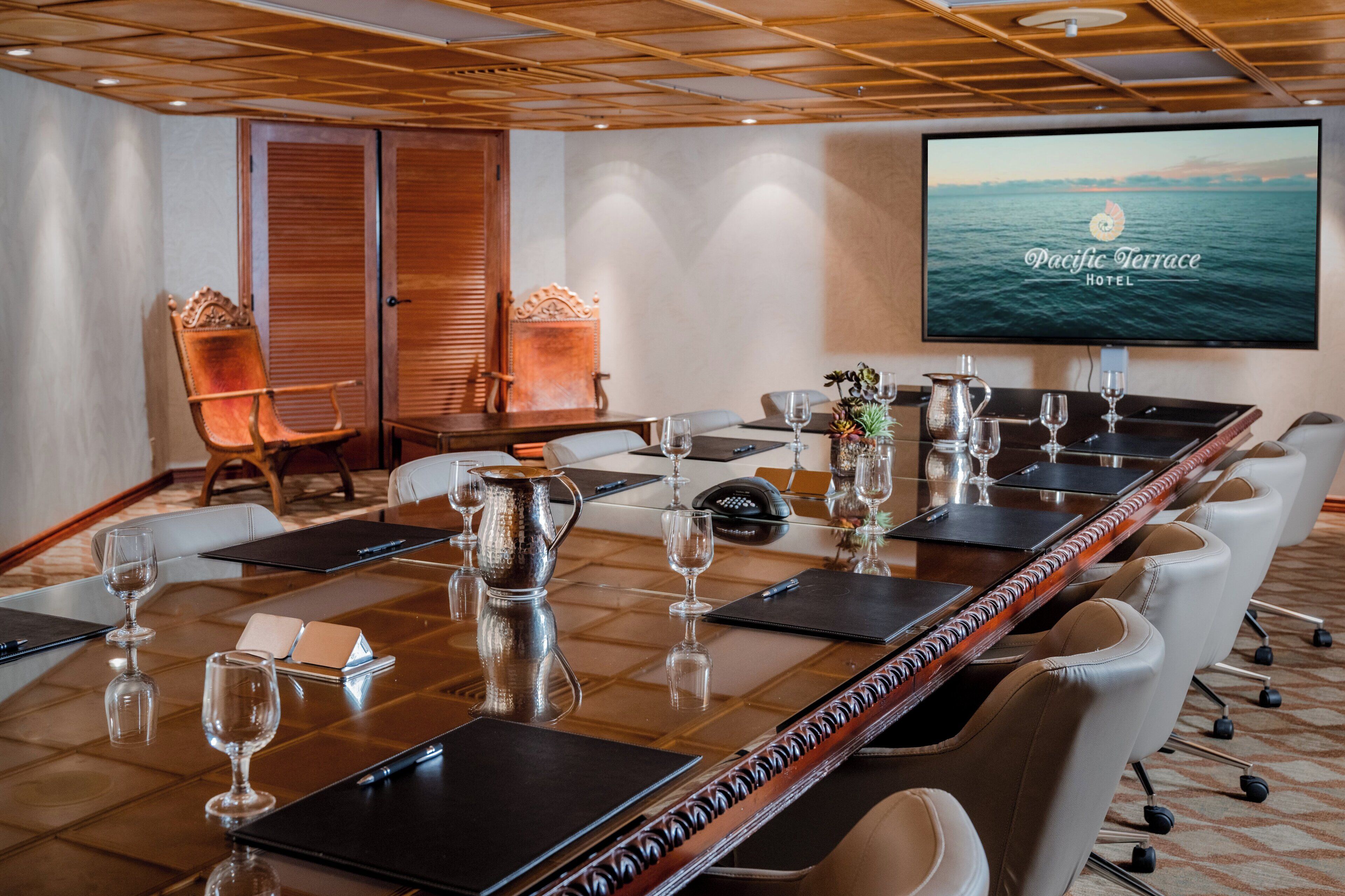 A conference room with a long table, chairs, a television screen displaying 'Pacific Terrace Hotel', and a decorative wall panel.