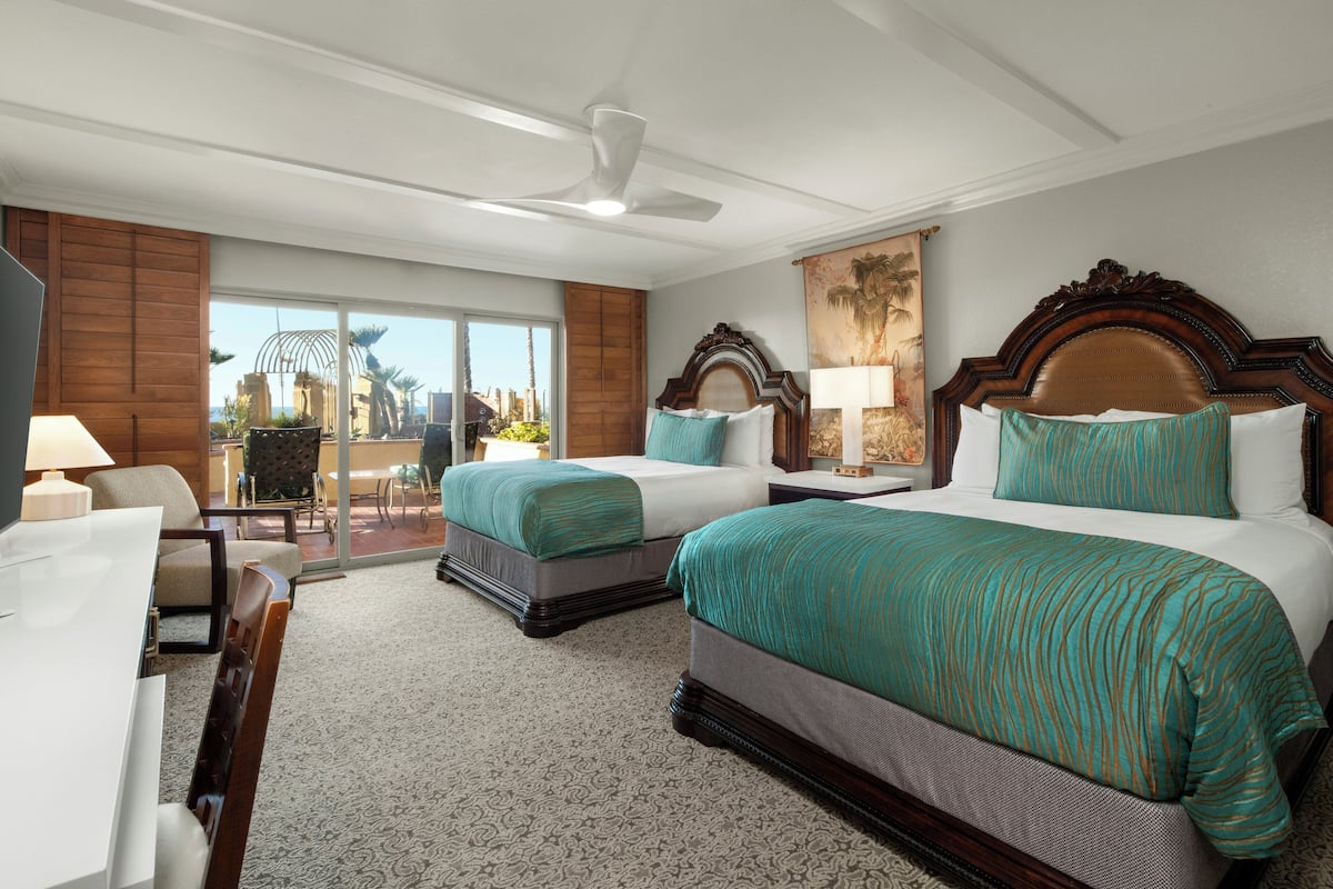A hotel room with two beds, a desk, and a chair. There is a balcony with a view of palm trees and buildings.