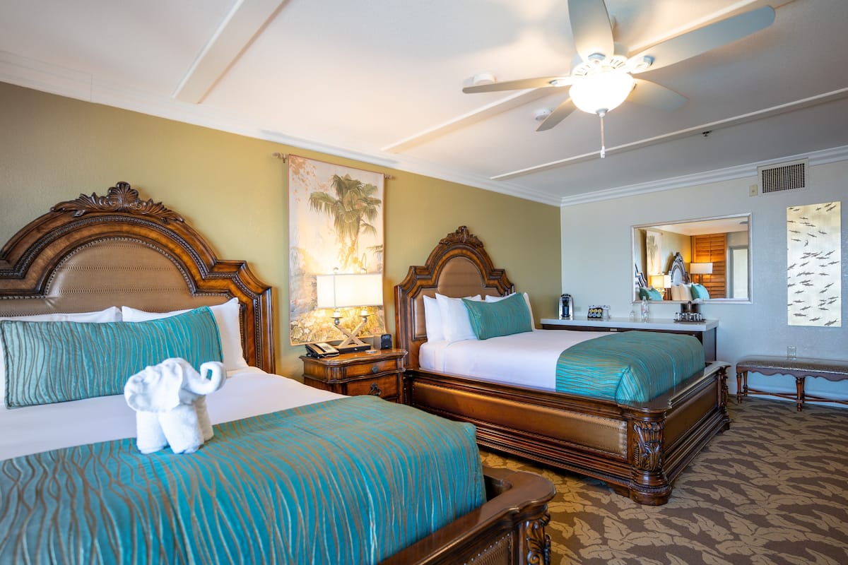 A hotel room with two beds, a ceiling fan, a nightstand with a lamp, and a painting on the wall.