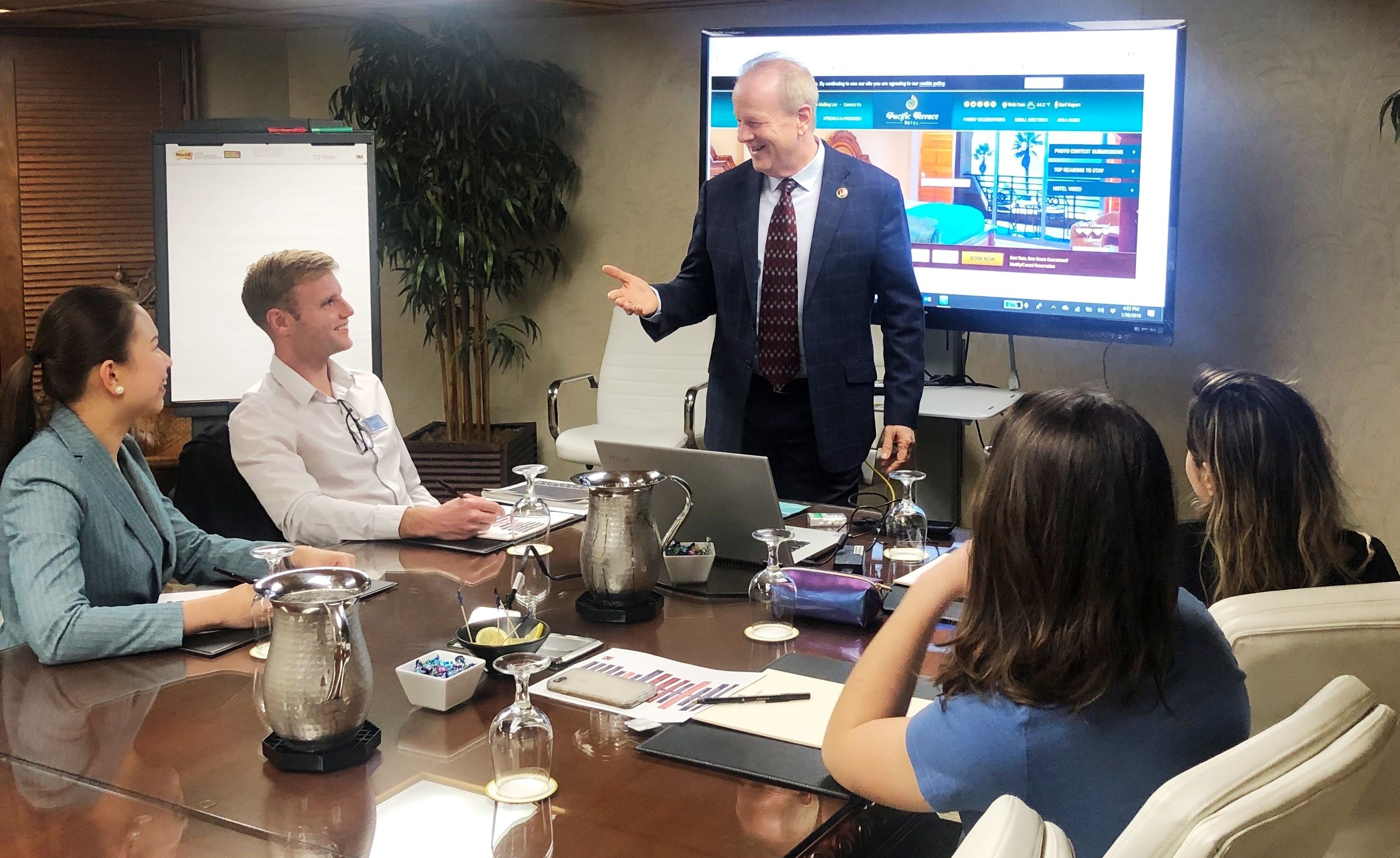 A business meeting in a conference room with a presentation screen, a whiteboard, and a laptop.