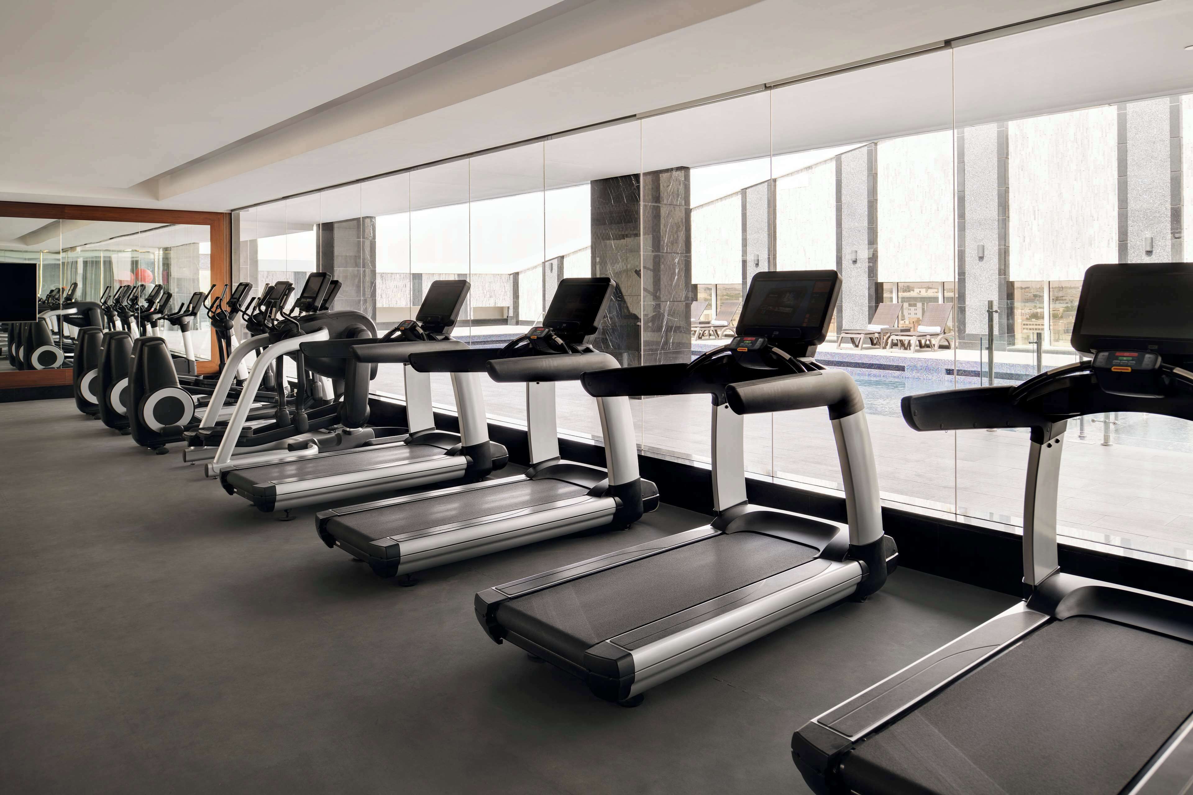 A row of stationary bikes and treadmills in a gym with large windows and a view of a pool.