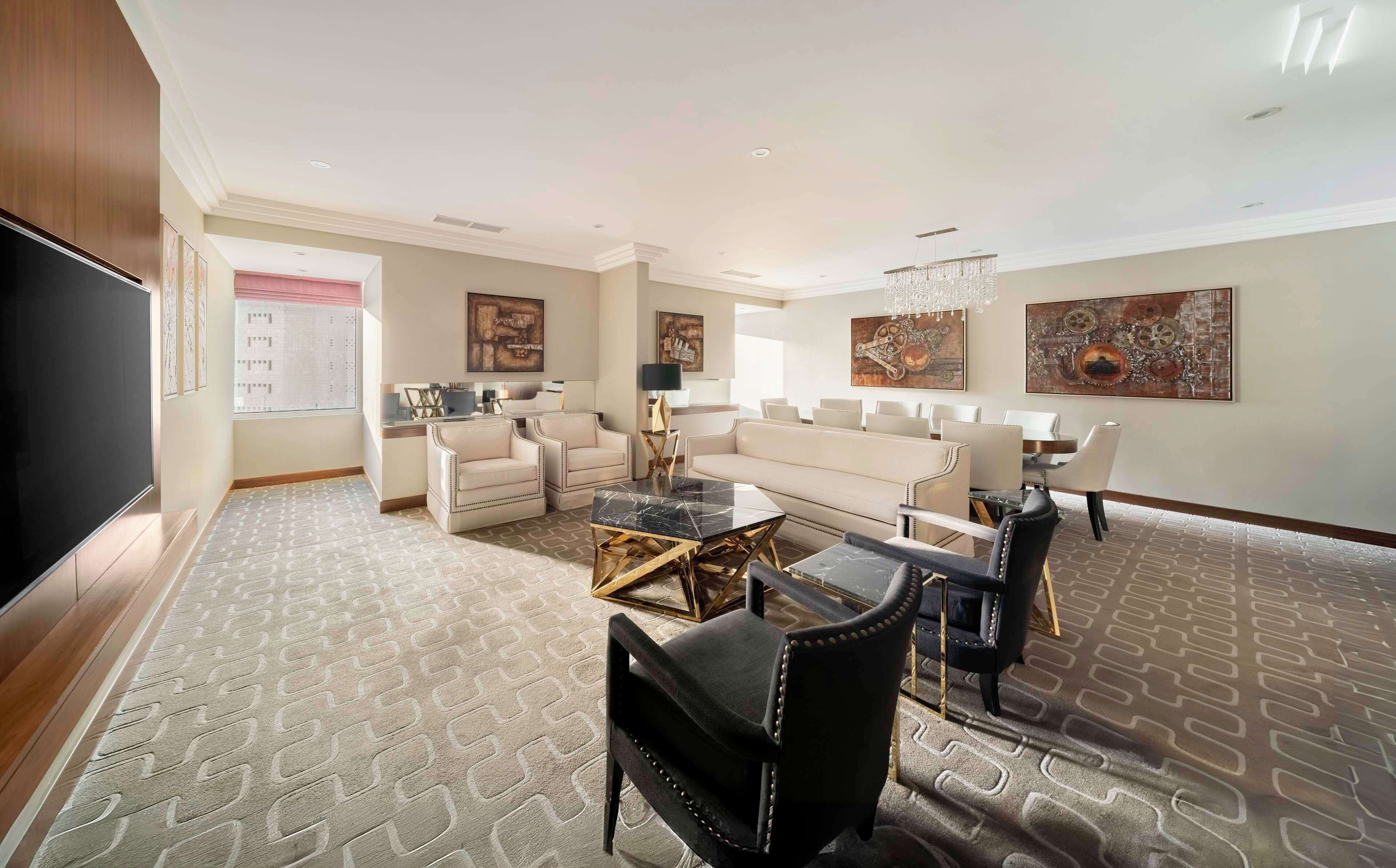 A spacious living room with a large flat-screen TV, a sofa set, a dining area with chairs, and a patterned carpet.