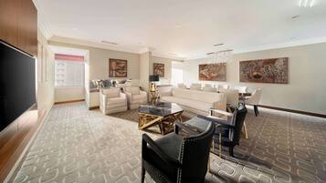 A spacious living room with a large flat-screen TV, a sofa set, a dining area with chairs, and a patterned carpet.