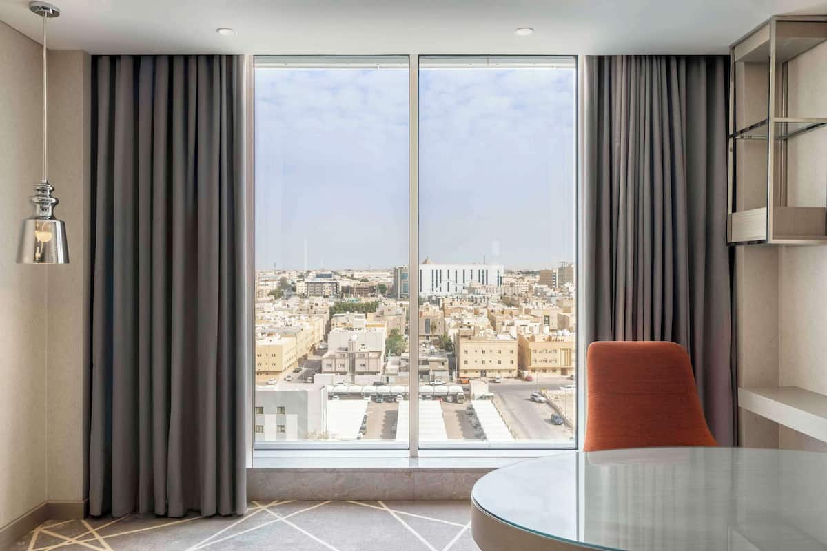 A hotel room with a large window, a glass table, a red chair, and a hanging light fixture.