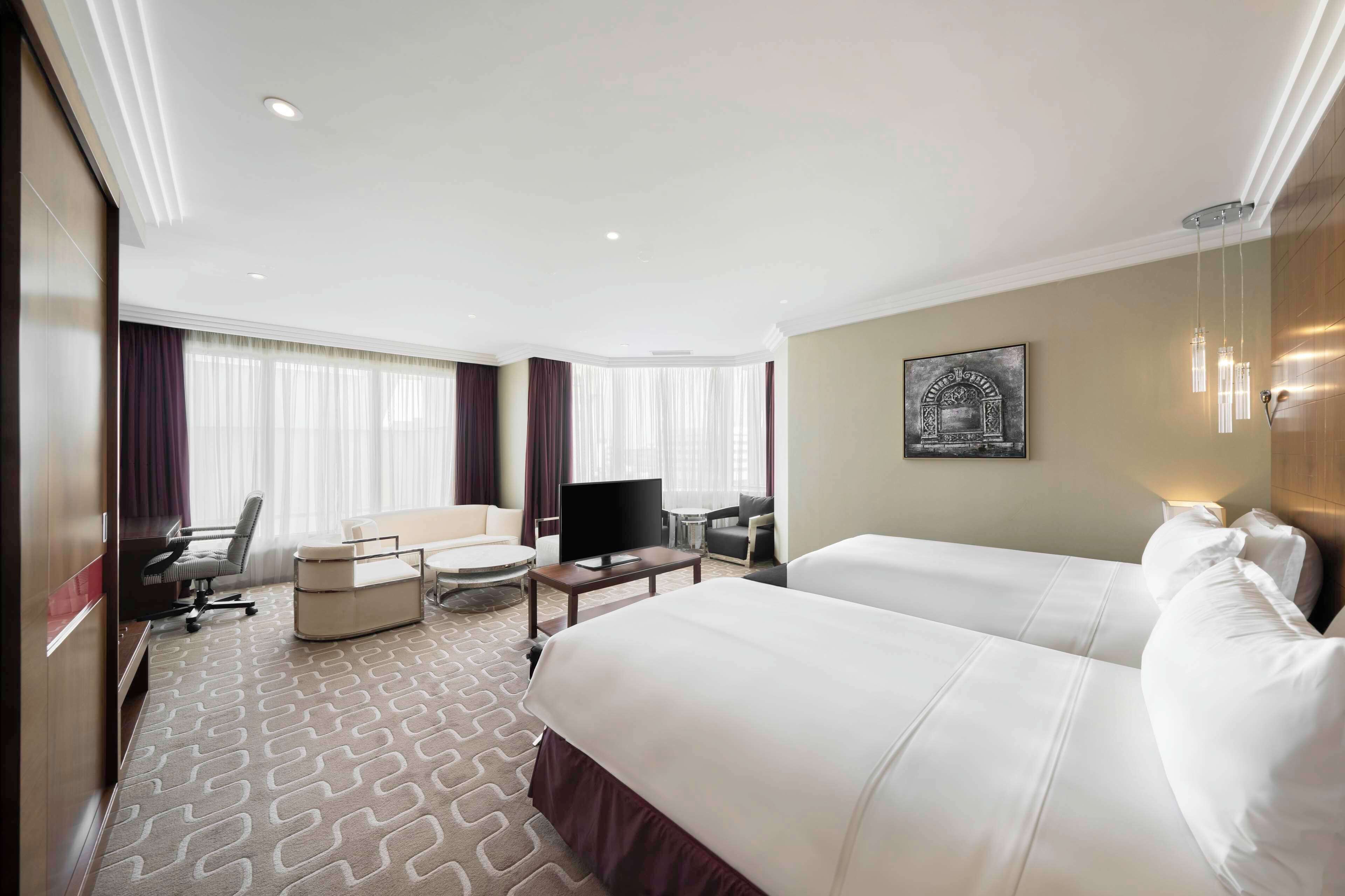 A hotel room with two beds, a desk with a chair, a television, and a large window with curtains.