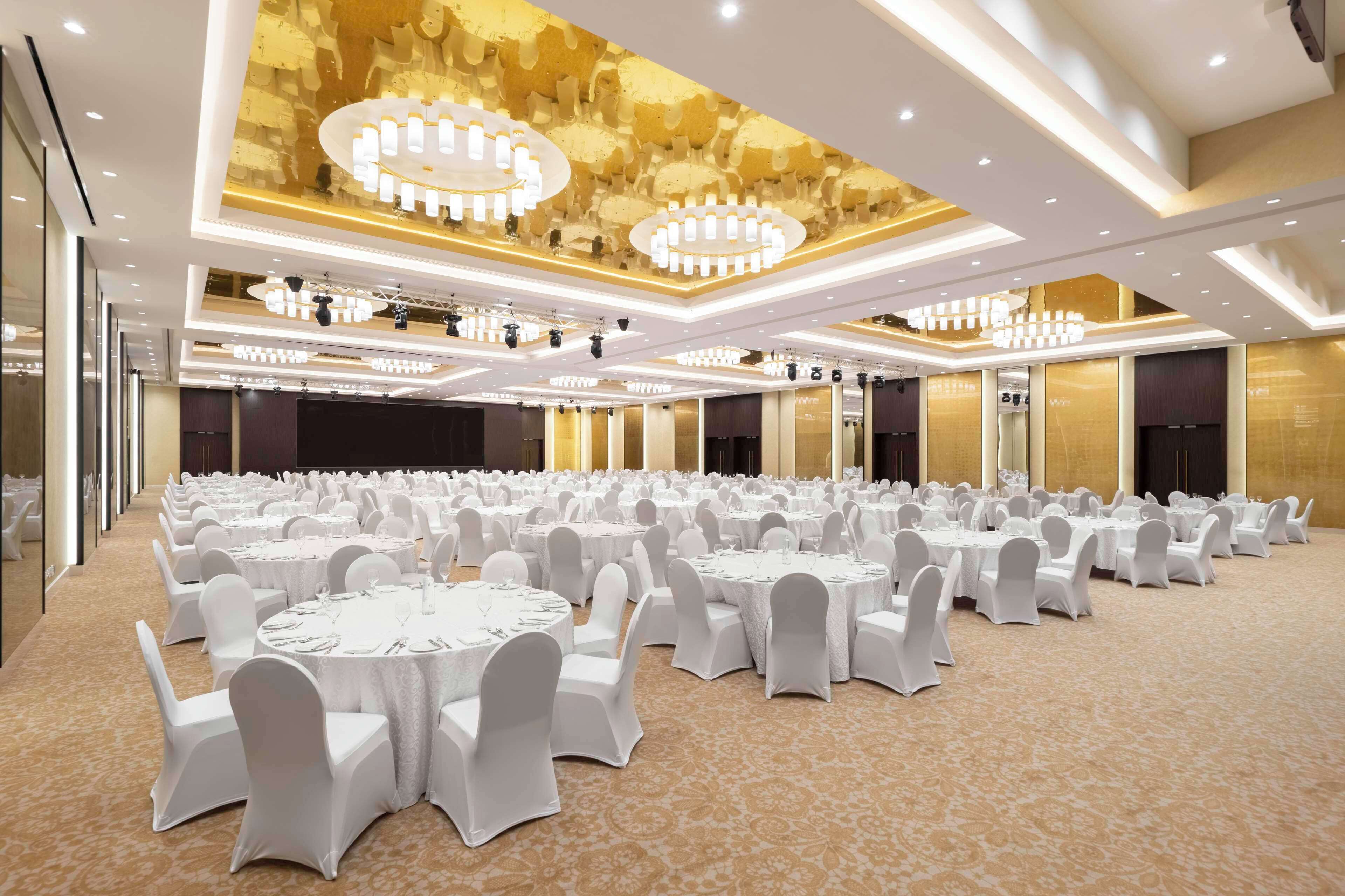 A large banquet hall with round tables and white chairs, featuring a patterned carpet and a decorative ceiling with chandeliers.