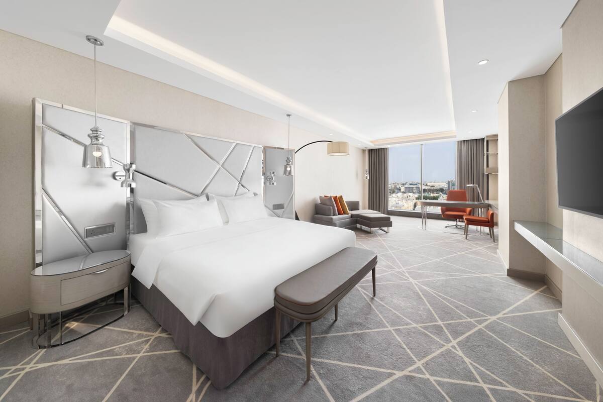 A modern hotel room with a large bed, a desk, a chair, and a TV.