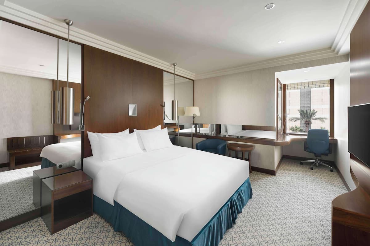 A hotel room with a large bed, a desk, a chair, a TV, and a window with a view.
