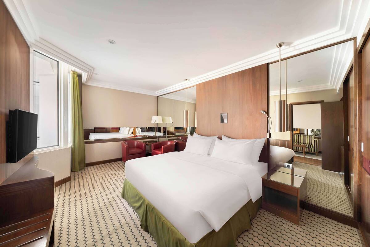 A hotel room with a large bed, a desk with a lamp, a TV, a mirror, and a red chair.