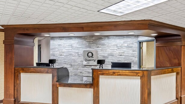 Un mostrador de recepción con un fondo de pared de piedra, un cartel que dice 'Quality Choice Hotels' y una zona de recepción con un mostrador de madera y dos sillas.