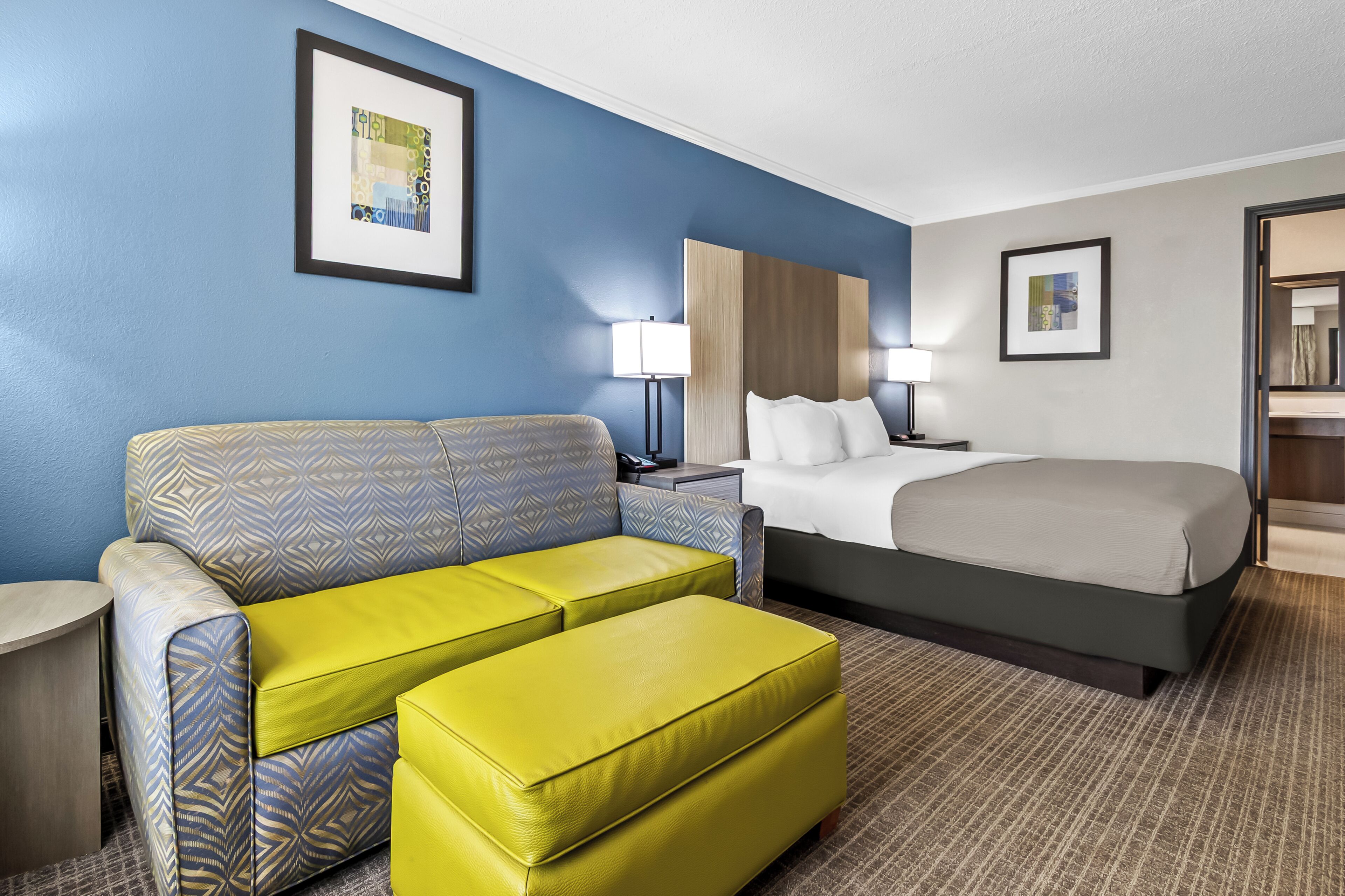 Ein Hotelzimmer mit Bett, Sofa, Hocker, Nachttisch, Lampe und zwei gerahmten Bildern an der Wand.