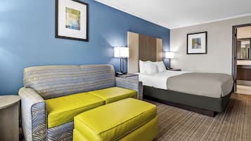 Ein Hotelzimmer mit Bett, Sofa, Hocker, Nachttisch, Lampe und zwei gerahmten Bildern an der Wand.