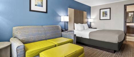 A hotel room with a bed, a sofa, an ottoman, a nightstand, a lamp, and two framed pictures on the wall.
