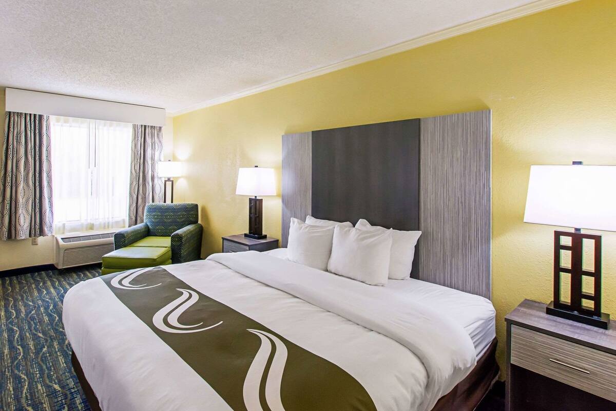 Quarto de hotel com uma cama grande, duas mesas de cabeceira com luminárias, uma cadeira verde e uma janela com cortinas.