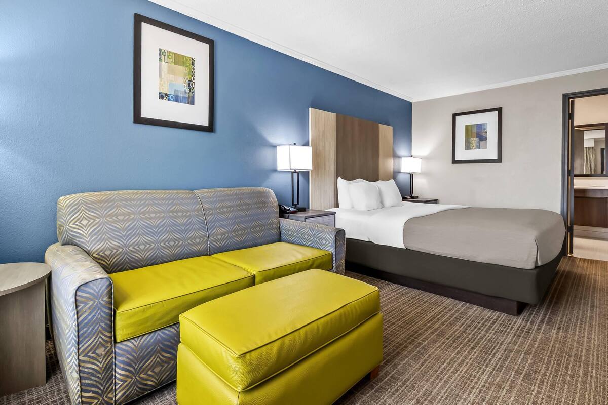 A hotel room with a bed, a sofa, an ottoman, a nightstand, and a lamp.