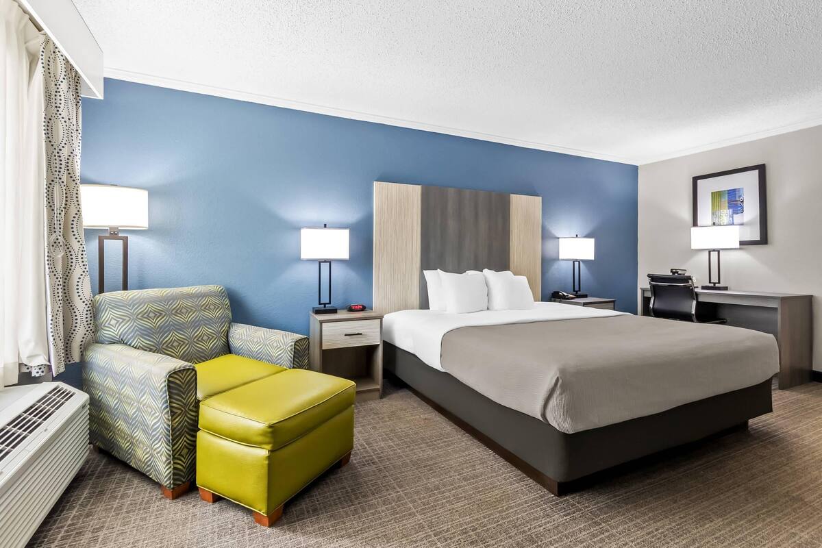 A hotel room with a bed, a desk, a chair, a lamp, and a radiator.