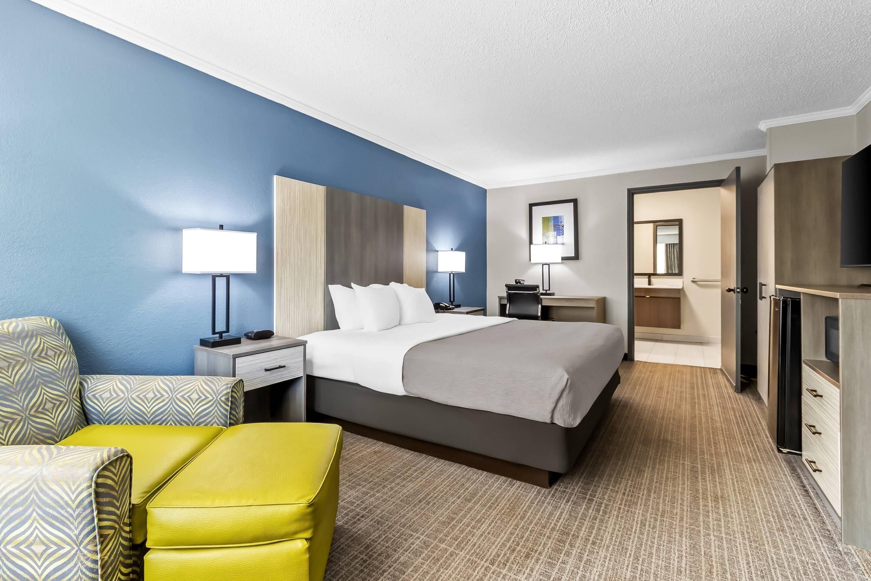 A hotel room with a bed, a desk, a chair, a TV, and a bathroom visible through an open door.