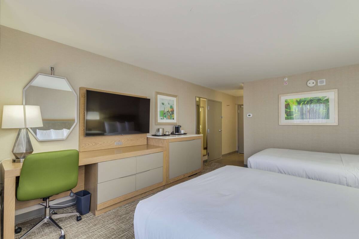 A hotel room with a desk, a green chair, a bed, a TV, and a painting on the wall.