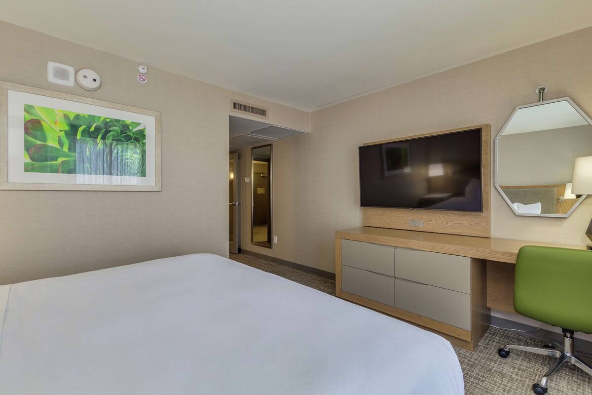 A hotel room with a large bed, a desk with a flat-screen TV, a green office chair, and a mirror on the wall.