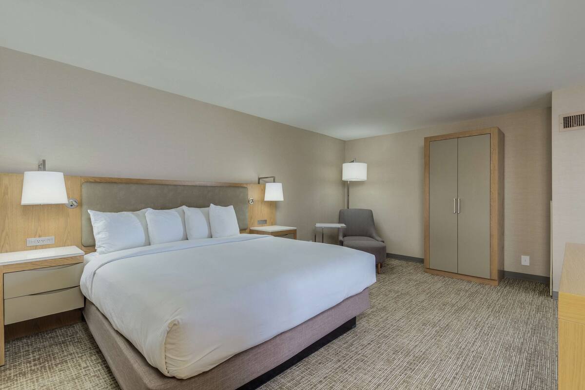 A hotel room with a large bed, two bedside tables with lamps, a chair, a wardrobe, and a carpeted floor.