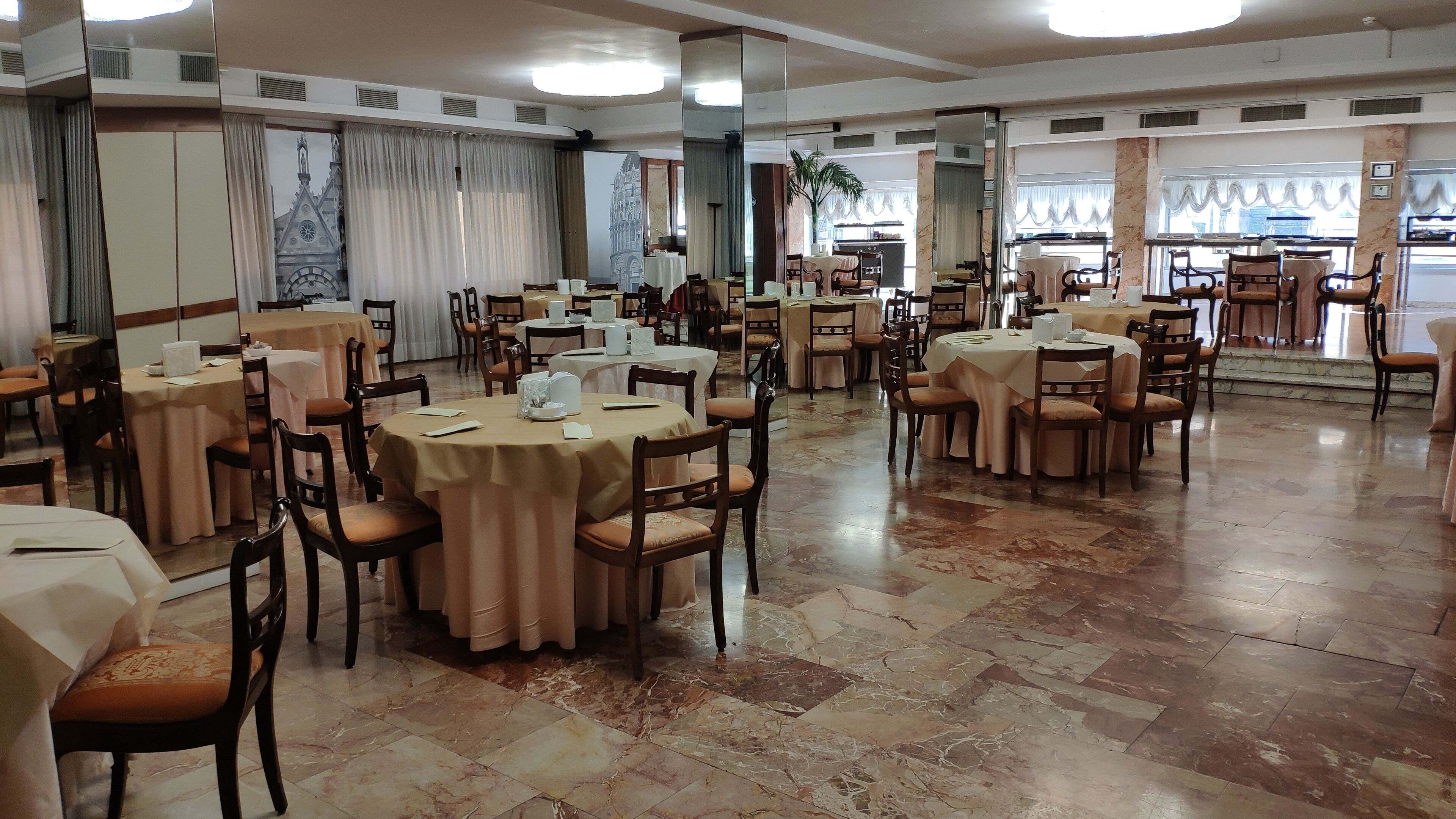 A spacious dining area with multiple round tables and chairs, marble flooring, and large windows with curtains.