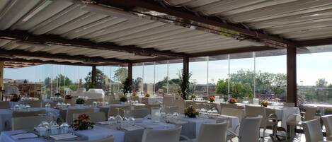 Un espacio al aire libre techado con mesas dispuestas para una comida, ventanas amplias y vistas del paisaje circundante.