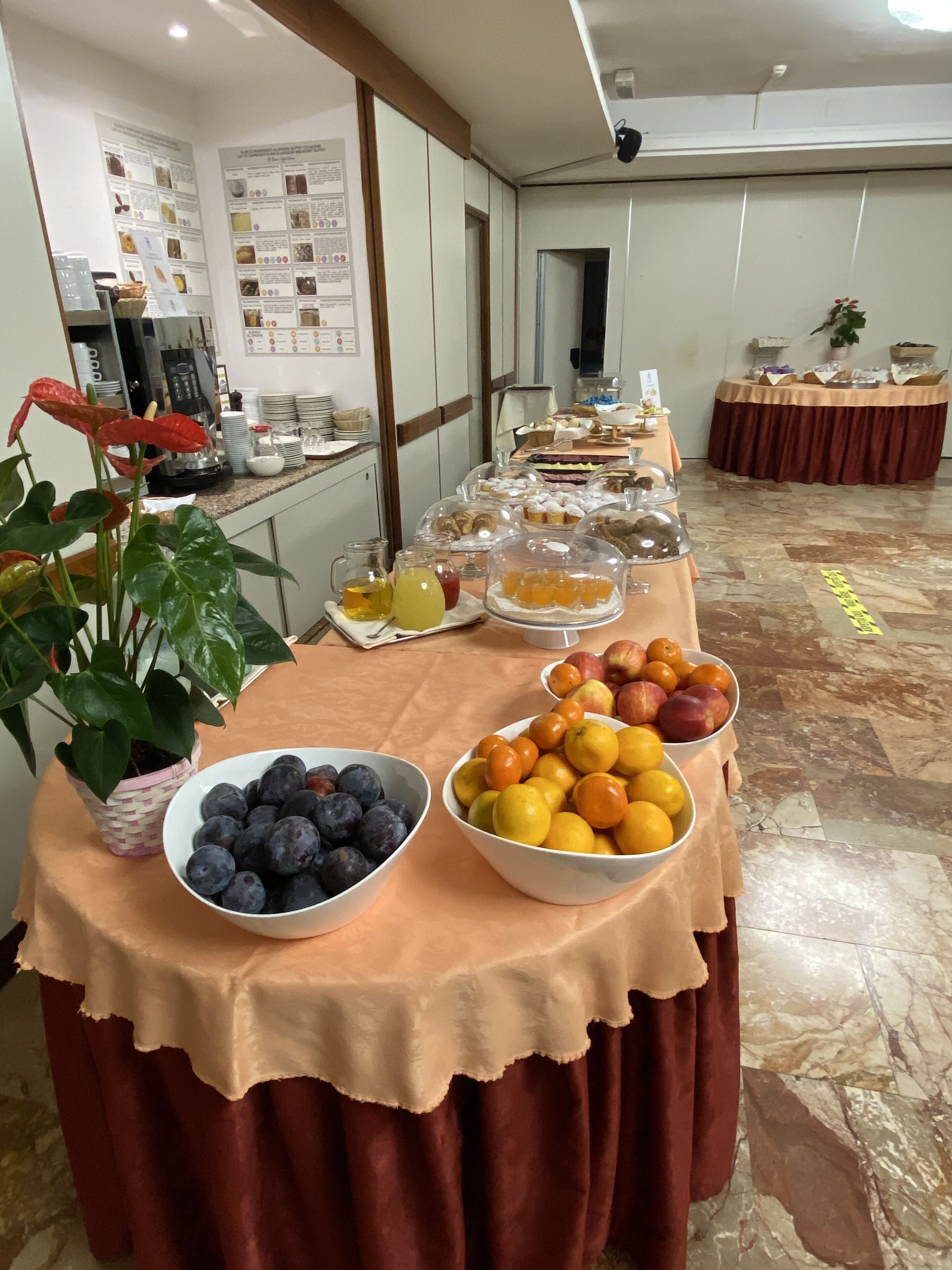 A buffet table with various fruits and pastries, a coffee machine, and informational posters on the wall.