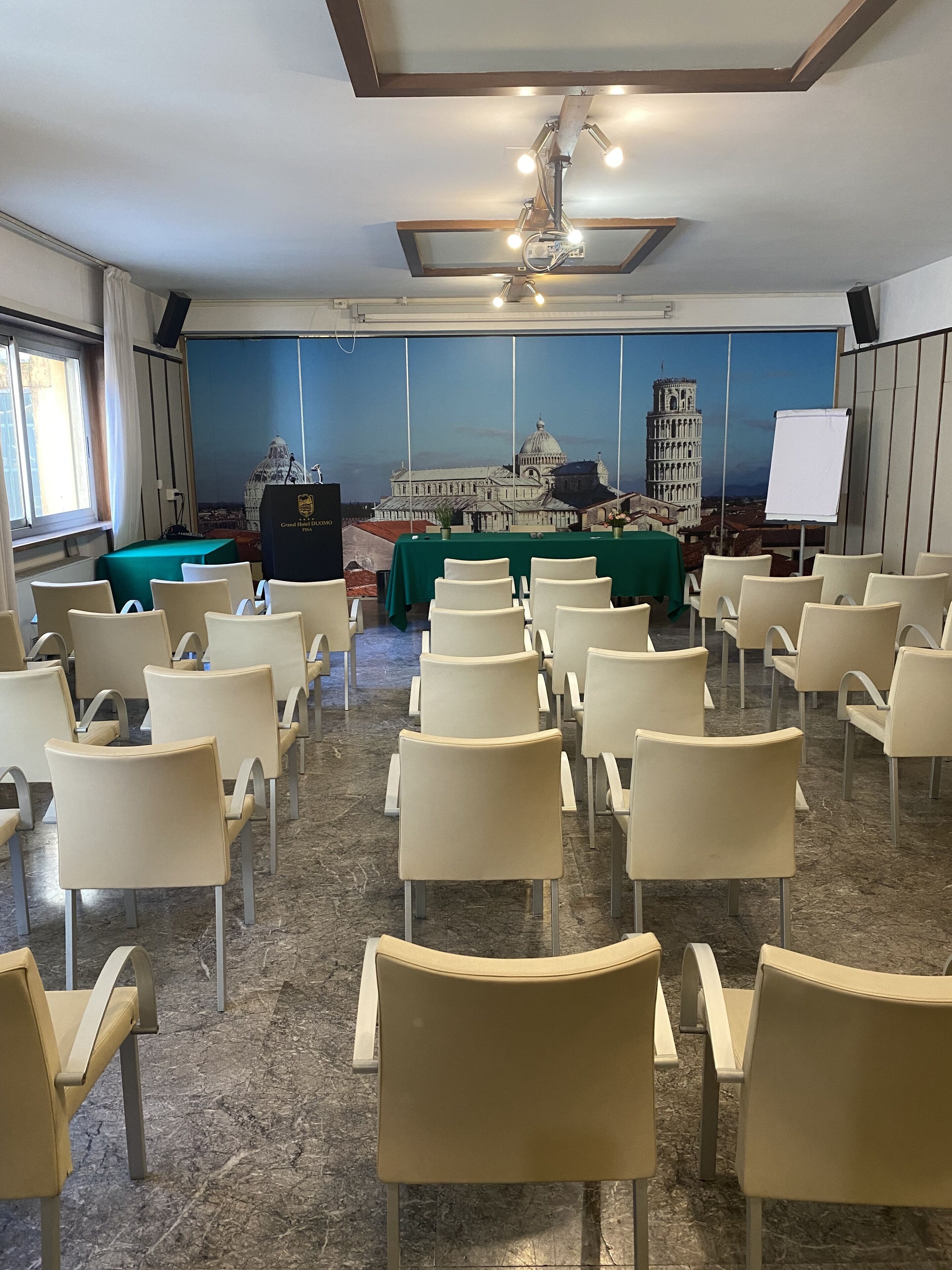 A conference room with rows of chairs, a green table, a podium, and a backdrop of the Leaning Tower of Pisa.