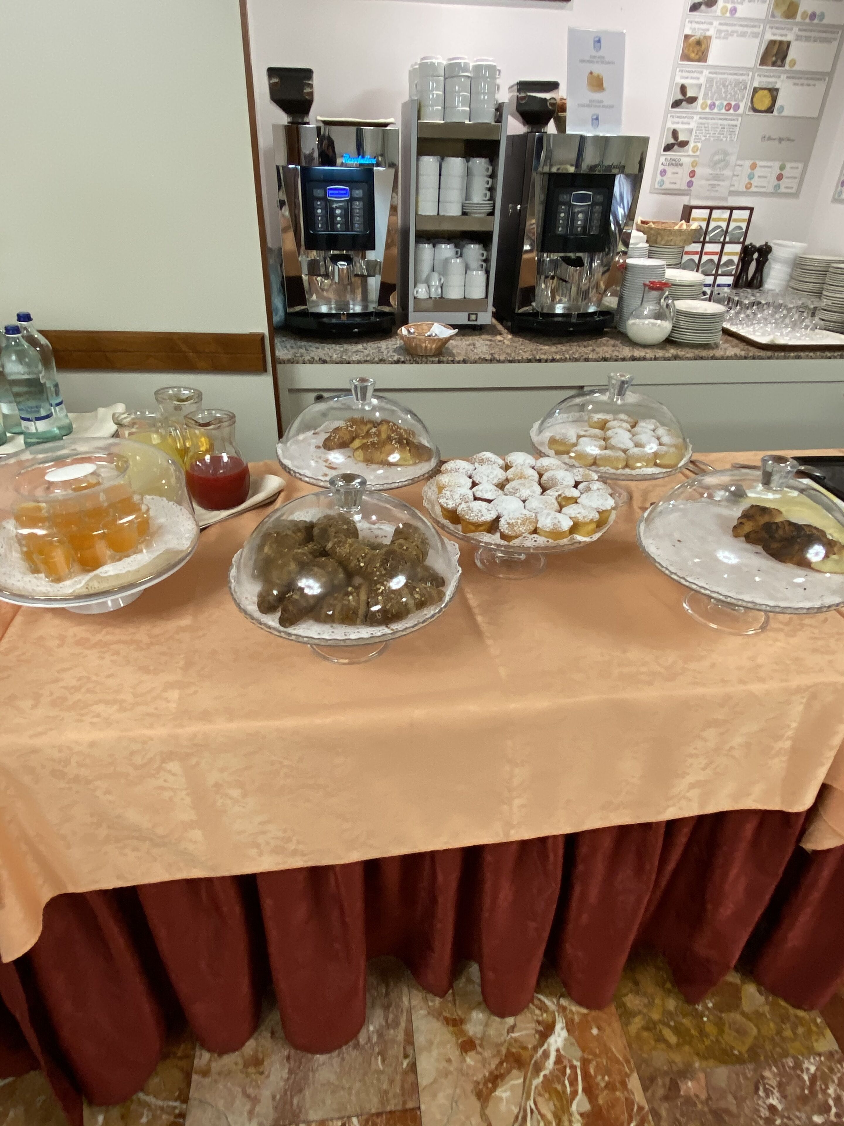 Un tavolo del buffet con diversi alimenti sotto cupole di vetro, un angolo caffè con diverse macchinette e una parete con poster informativi.