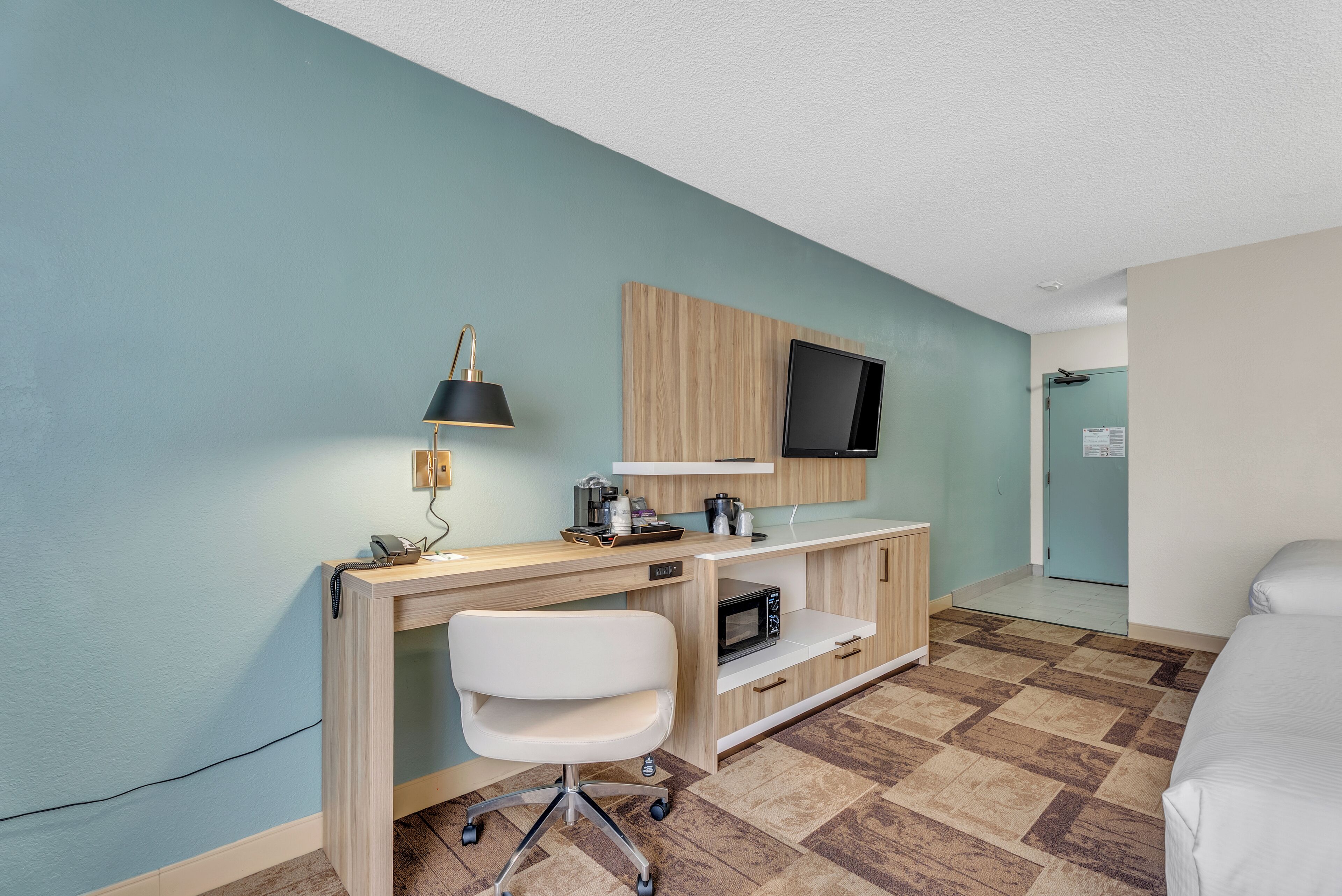 Une chambre d’hôtel avec un bureau en bois, une chaise blanche, un téléviseur à écran plat fixé au mur, un four à micro-ondes et un lit.