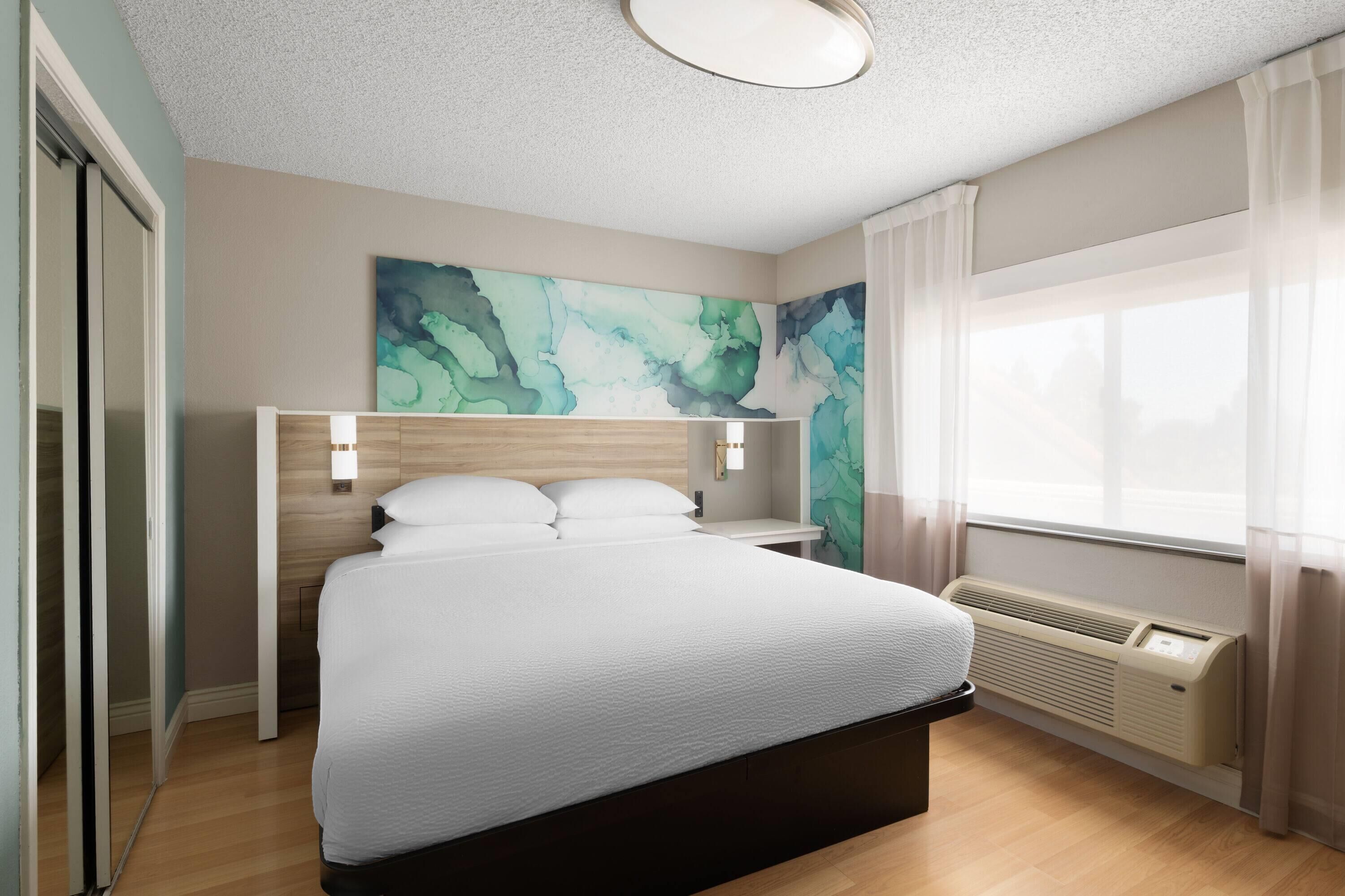 Quarto de hotel moderno com uma cama grande, mesa de cabeceira, luminária na parede e uma janela com cortinas leves.