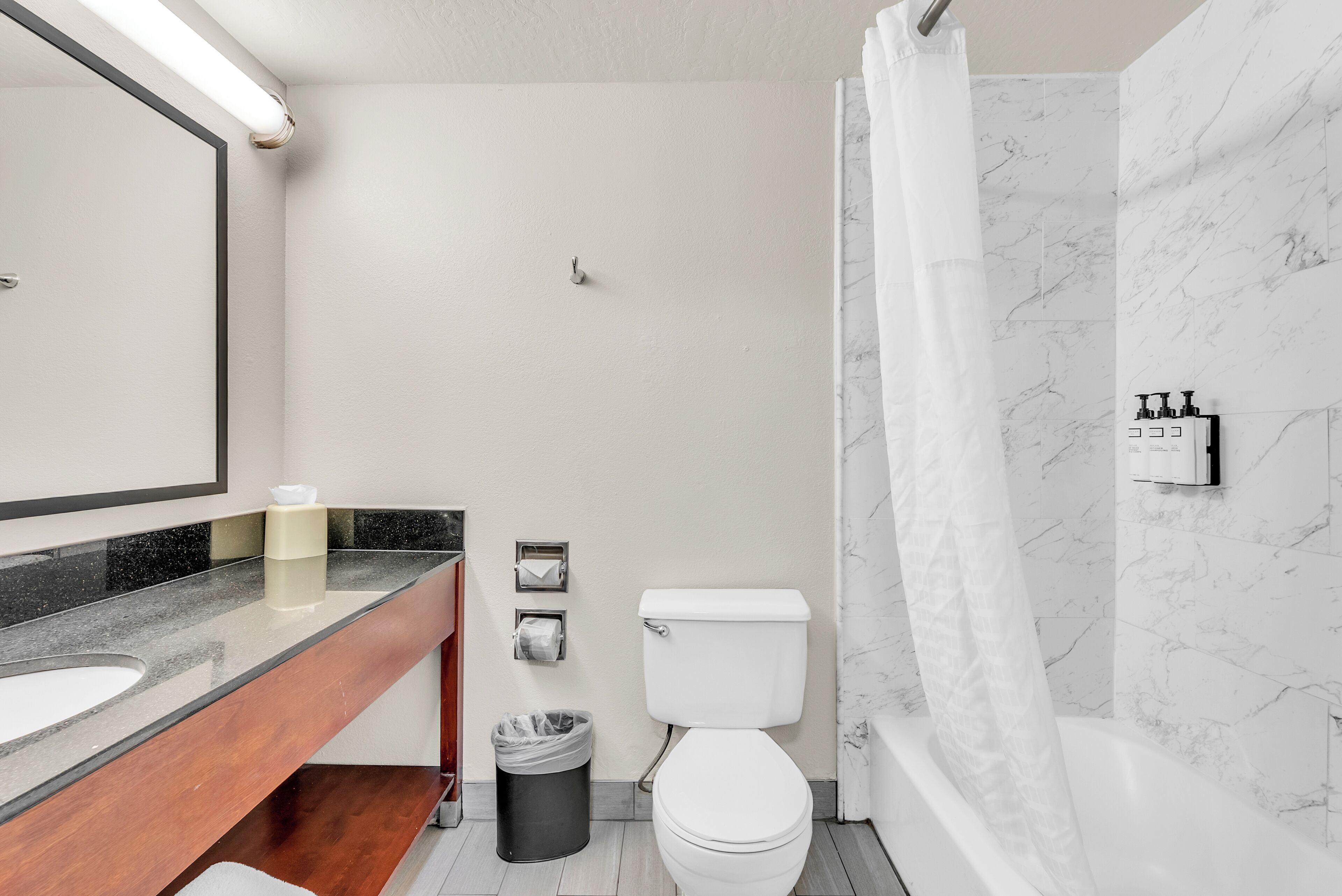 Un baño moderno con pared de mármol, inodoro blanco, tocador de madera, espejo y cortina de ducha.