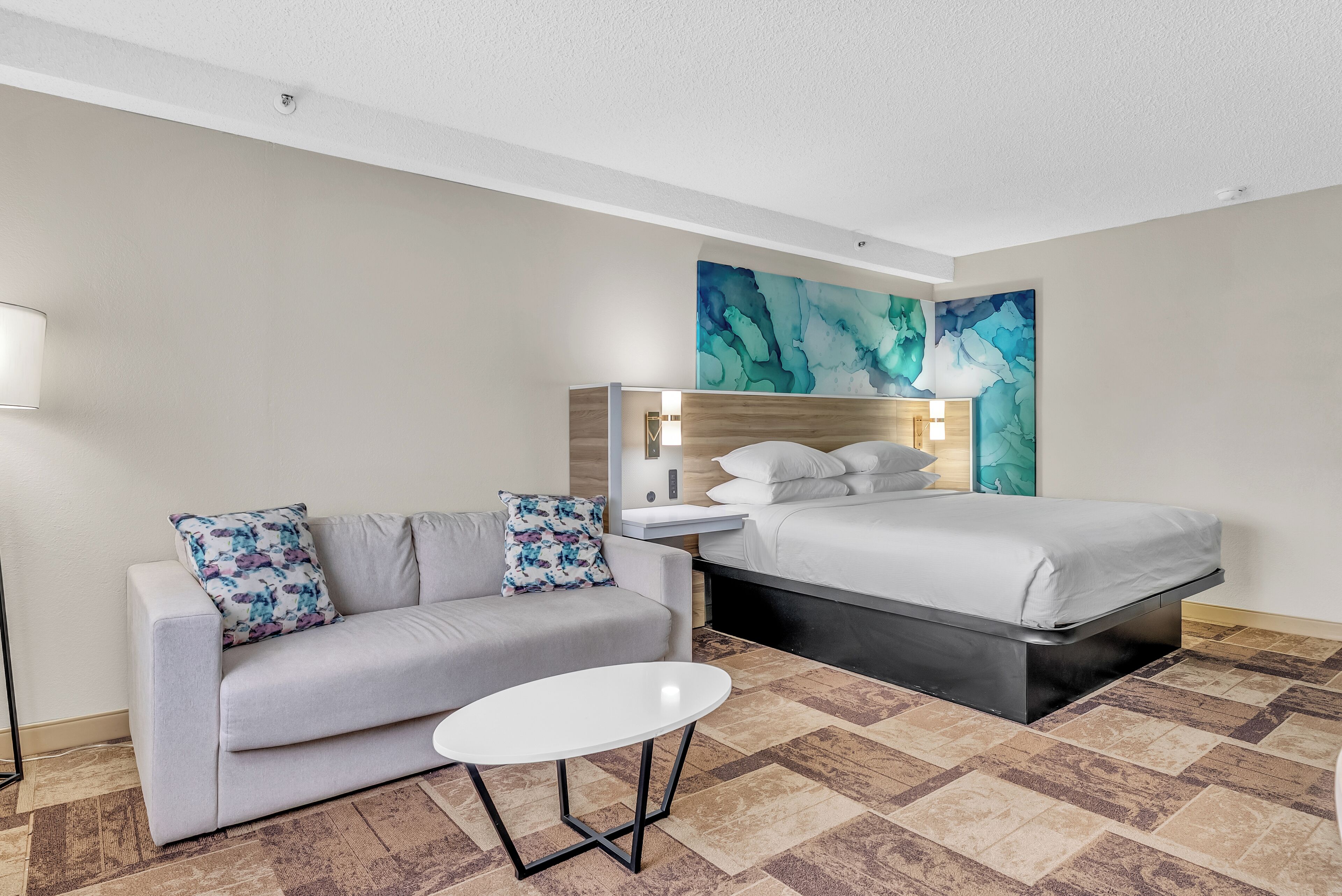 A hotel room with a sofa, a small round table, a bed, and a large abstract painting on the wall.