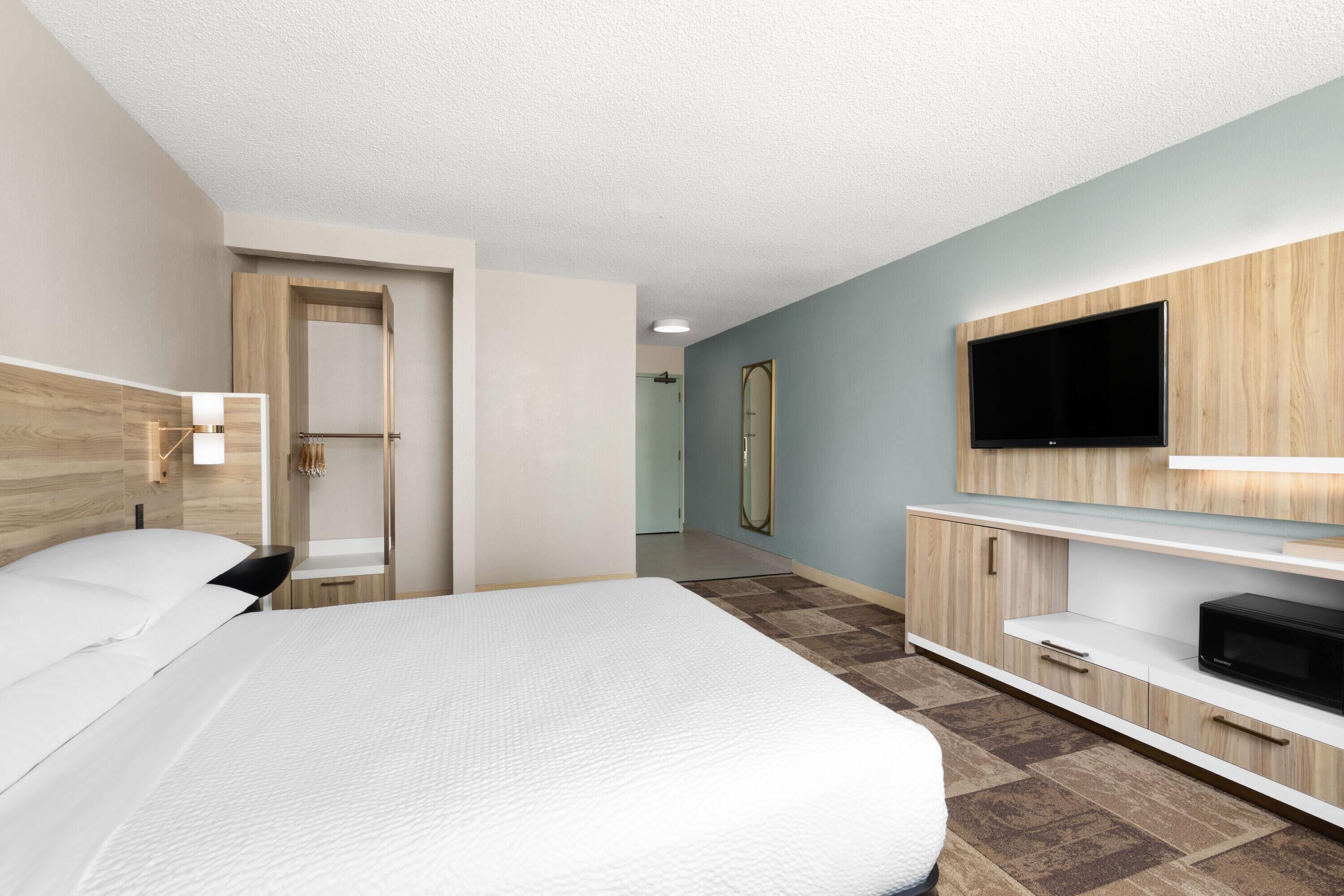 A modern hotel room with a large bed, a TV mounted on the wall, a wooden cabinet, and a mirror.