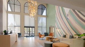 Een ruime lobby met een receptiebalie, comfortabele zitplaatsen, grote ramen en een kleurrijke abstracte muurschildering.
