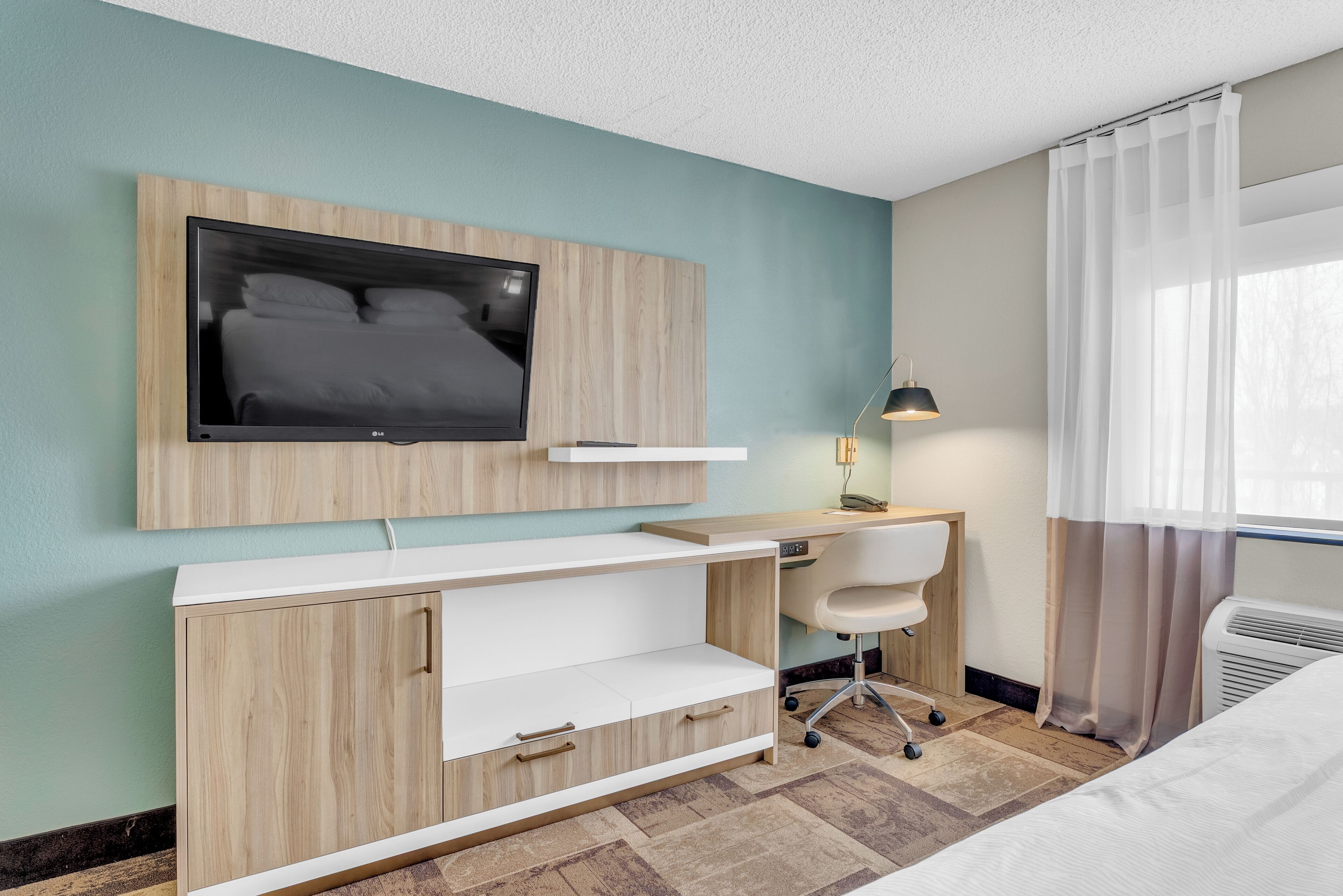 Una habitación de hotel moderna con un escritorio de madera, un televisor de pantalla plana montado en la pared, una silla blanca y una cama con ropa de cama blanca.