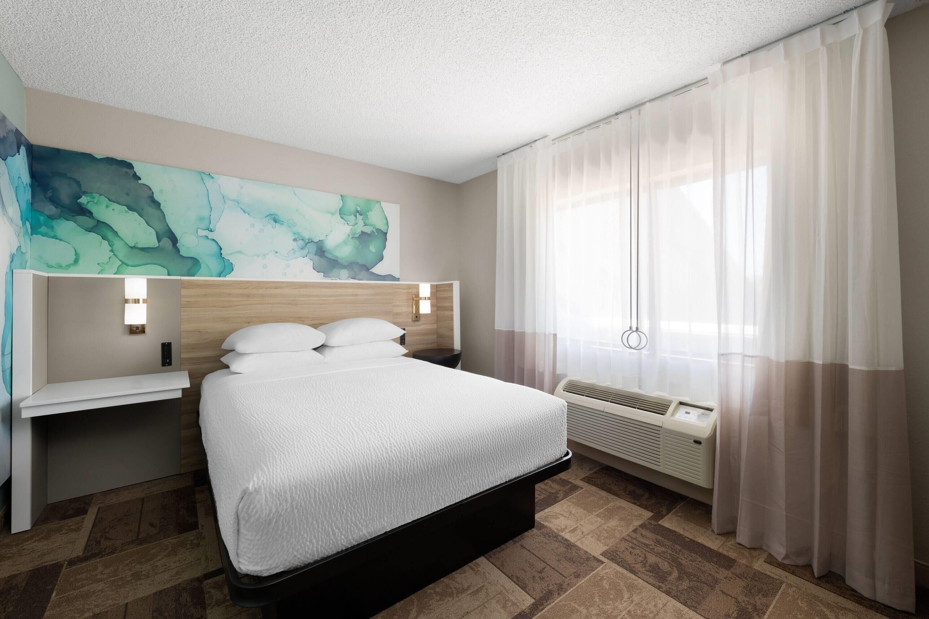 A hotel room with a bed, a nightstand, a wall-mounted light, a window with sheer curtains, and a wall art piece.