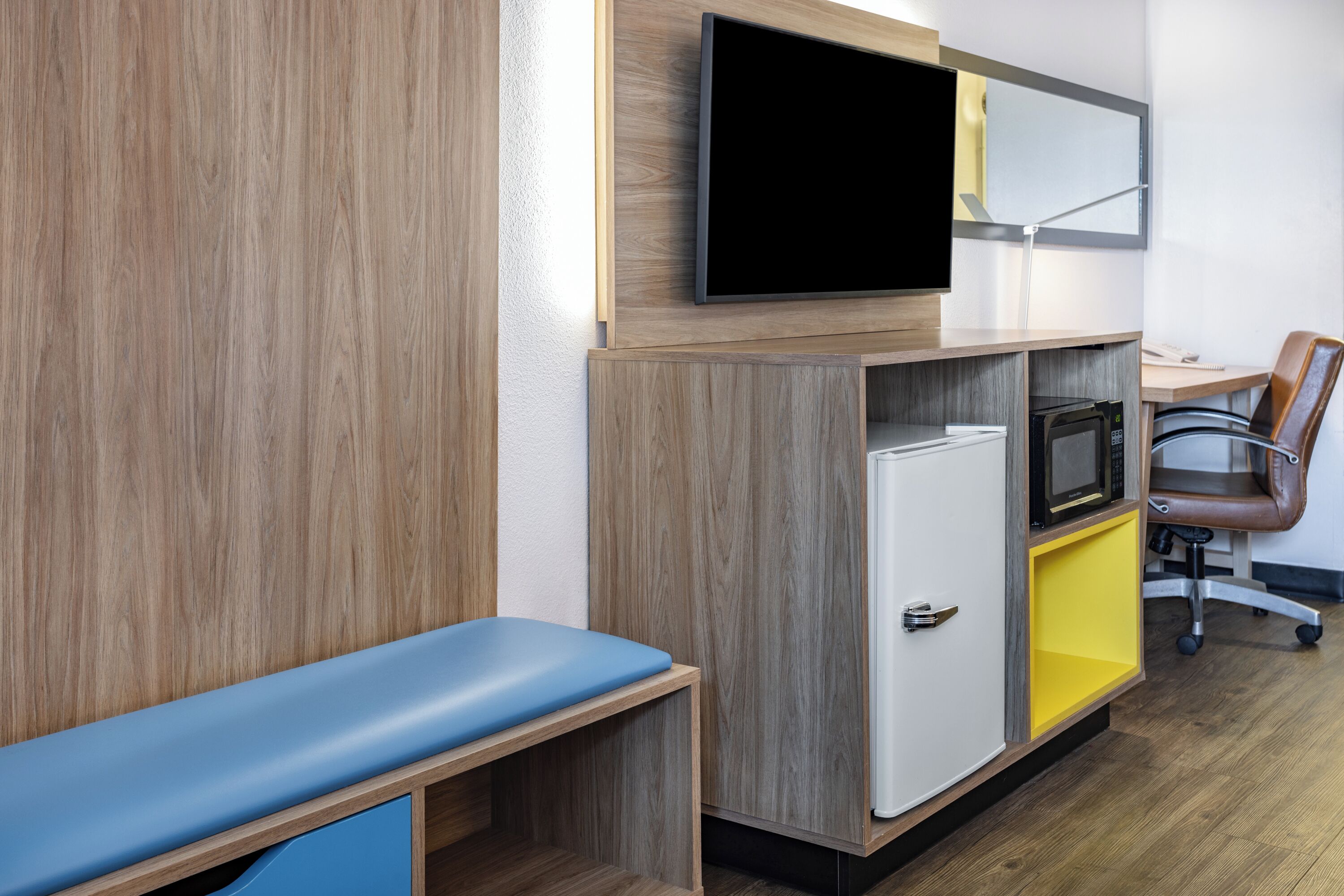 Een moderne hotelkamer met een houten wand, een flatscreen televisie boven een bureau, een magnetron, een minibar en een blauwe bank, plus een bruine leren fauteuil.
